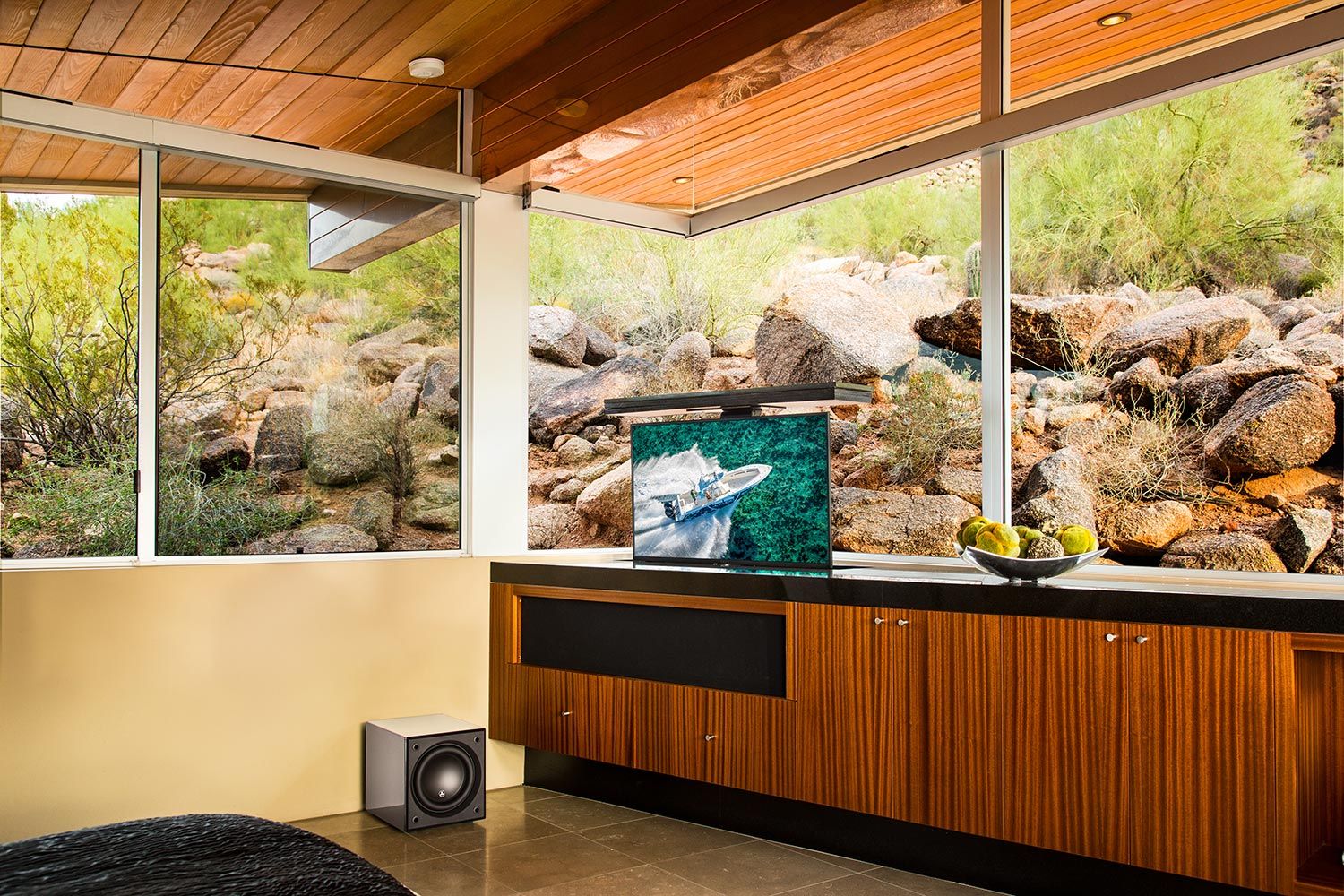 A stylish living room with large windows showcasing natural scenery. A sleek JL Audio Dominion® subwoofer is positioned in the corner, providing powerful, compact bass for an immersive audio experience.