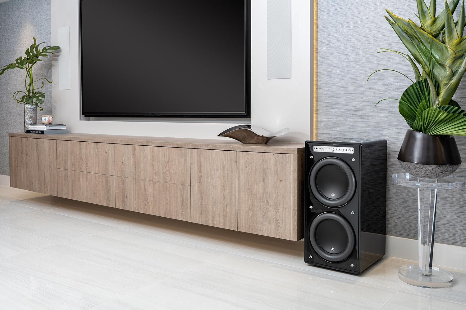 A modern living room with a wall-mounted TV and minimalist decor. A JL Audio Fathom® v2 subwoofer is placed next to the TV stand, delivering deep, accurate bass for an exceptional audio experience.